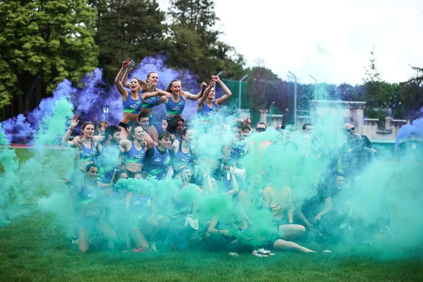 Athlètes du club en maillot bleu et vert qui célèbrent sur la pelouse, certaines portées sur les épaules, au milieu de fumigènes bleus et verts