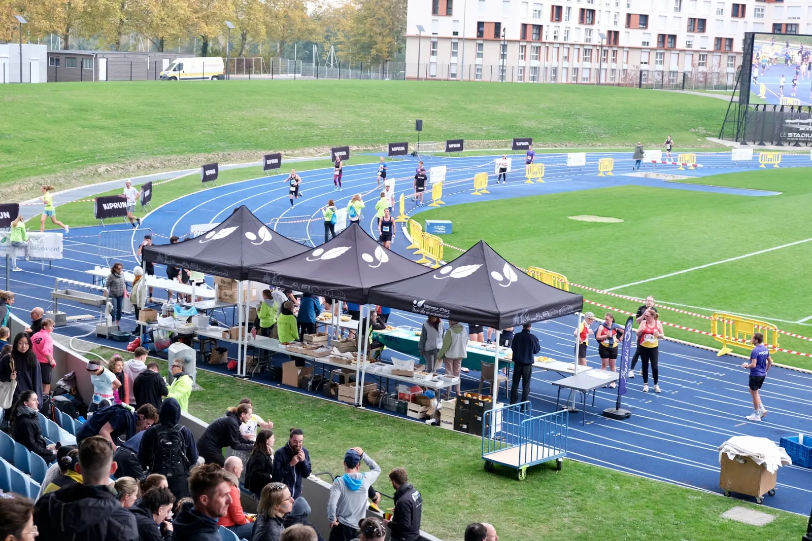Compétition sur une piste bleue : tentes noires des clubs, athlètes en course et public dans les gradins au premier plan