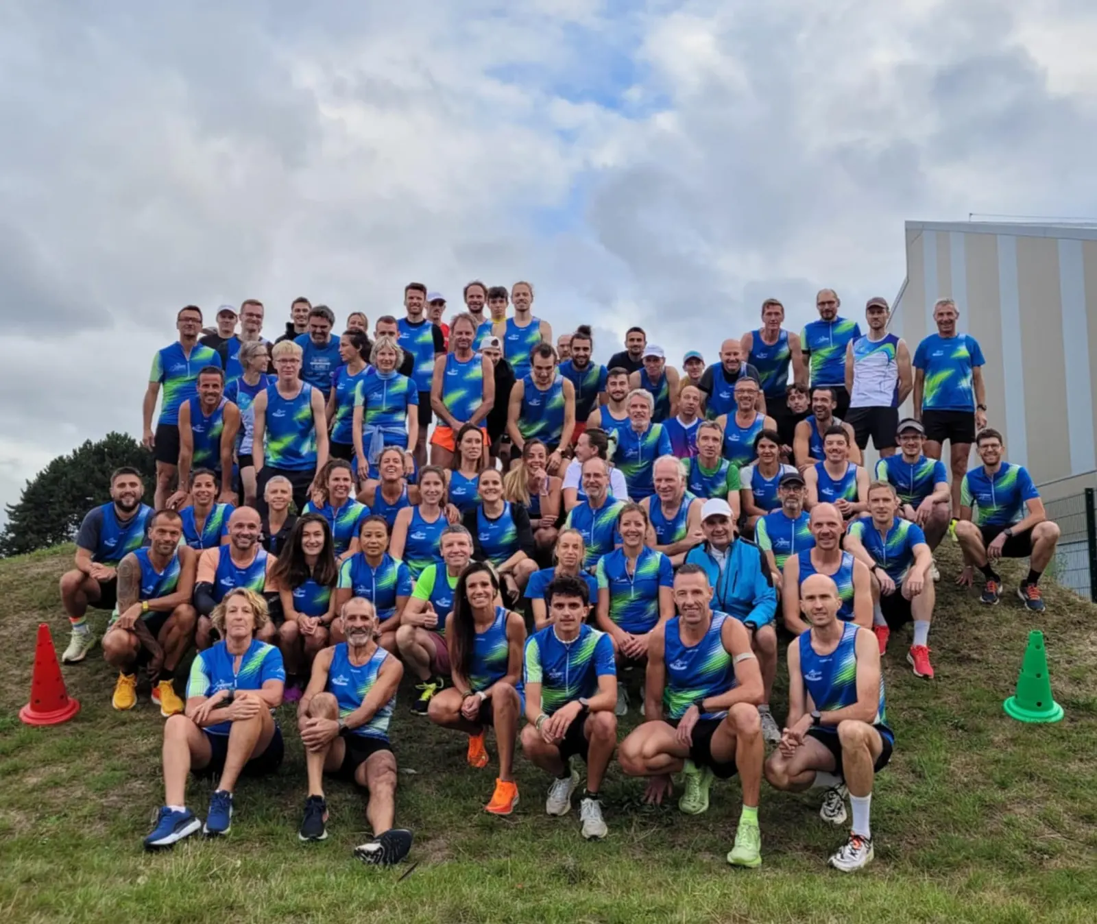 Photo de groupe d'une soixantaine de coureurs du club en maillot bleu et vert, assis et debout sur une butte herbeuse