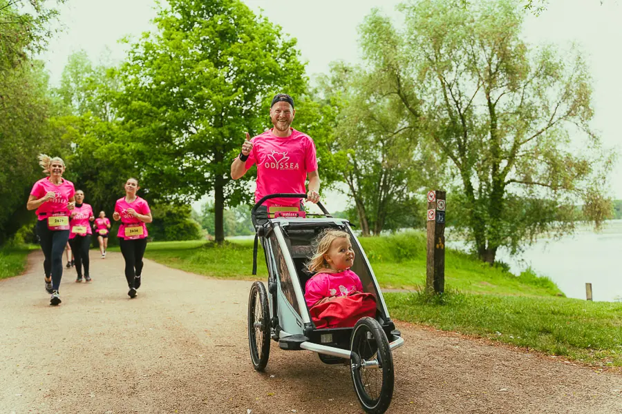 Coureurs en tee-shirt rose de la course Odyssea au bord d'un lac, dont un coureur qui pousse une petite fille dans une poussette de course